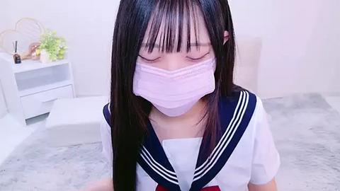 Media: A video of an Asian woman with long black hair, wearing a school uniform and a pink face mask, kneeling in a modern, minimalist room with white walls and a light grey floor.