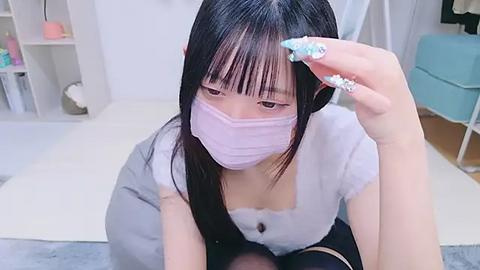 Media: Video of an Asian woman with long black hair, wearing a pink face mask, adjusting her hair in a stylish, modern room with white furniture and pastel decor.