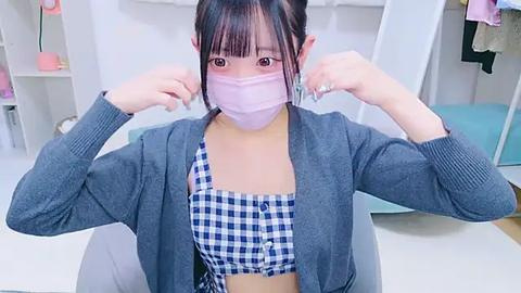 Media: Video of an East Asian woman in a light gray cardigan over a blue and white checkered crop top, adjusting a pink face mask. She has dark hair in pigtails. Background features a white wardrobe with hanging clothes.