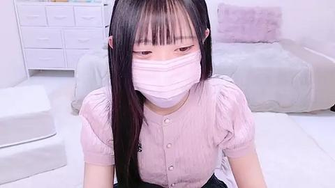 Media: A video of an East Asian woman with long black hair, wearing a pink blouse and face mask, sitting on a white bed in a minimalist bedroom with white furniture.