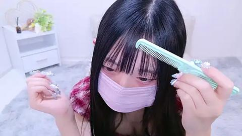 Media: Video of an Asian woman with long black hair, wearing a pink face mask, holding a green hairbrush, and a plastic comb, in a minimalist, white room.