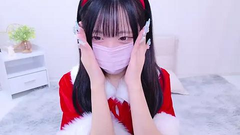 Media: Video of an East Asian woman with long black hair, wearing a red Santa hat, a pink face mask, and a red and white Santa outfit, seated on a white furry rug, in a minimalistic white room with a green plant.