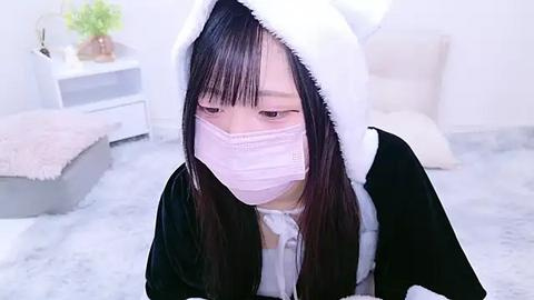 Media: Video of an Asian woman in a pink surgical mask and a white and black cat costume, sitting on a plush white carpet in a minimalist, light-colored room.