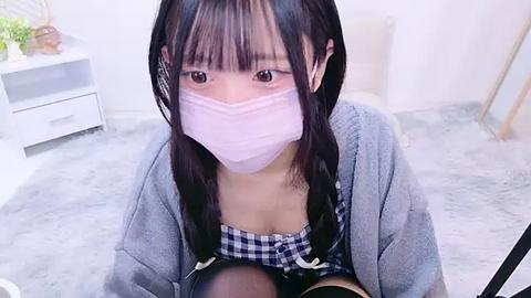 Media: Video of an Asian woman with long black hair, wearing a pink mask, plaid shirt, and gray cardigan, kneeling on a fluffy white rug in a minimalist room with white furniture.