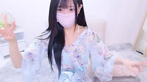 Media: Video of an Asian woman with long black hair, pale skin, and a light floral kimono, wearing a face mask, seated on a plush grey rug in a minimalist room with white walls and a small table.