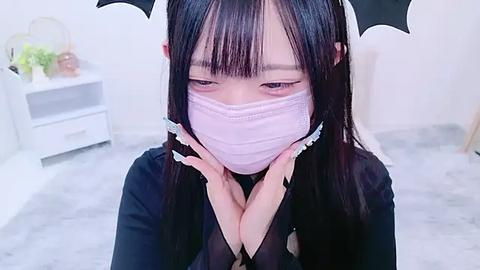 Media: A video of an Asian woman with long black hair, wearing a pink face mask and black bat headband, sitting on a grey carpeted floor in a minimalist room.