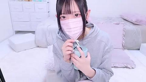 Media: A video of an Asian woman with long black hair and pale skin, wearing a pink face mask, grey hoodie, and holding a blue cup, sitting on a white floor in a minimalist bedroom with white furniture.