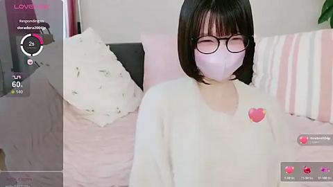 Media: Video of a young woman with short black hair and glasses, wearing a pink mask and white shirt, lying on a bed with pink and white pillows.