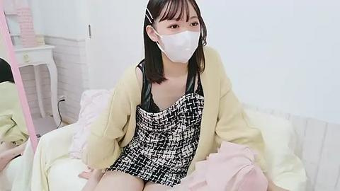 Media: Video of an Asian woman with straight black hair, wearing a white mask, black and white houndstooth dress, beige cardigan, sitting on a bed in a bright, minimalistic room.