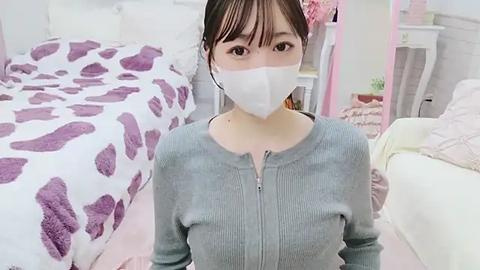 Media: Video of an East Asian woman with pale skin, wearing a white face mask, gray ribbed sweater, and dark hair, sitting on a pink carpet in a pastel-colored bedroom with white furniture and purple patterned bedding.