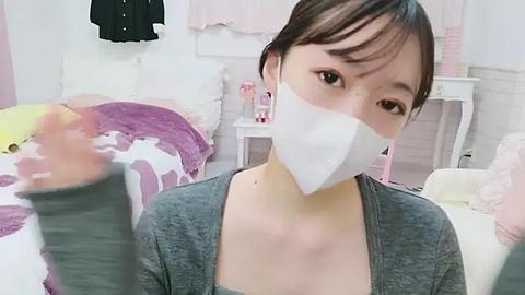 Media: A video of an East Asian woman with short black hair, wearing a grey cardigan and a white face mask, posing in a soft, pink-toned room with plush toys and pastel decor.