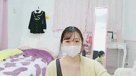 Media: Video of a young Asian woman in a pastel bedroom wearing a white face mask, black jacket, and yellow cardigan, sitting on a bed with a white and purple duvet.