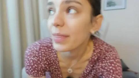 Media: Video of a woman with dark hair, fair skin, and brown eyes, wearing a maroon floral dress, gazing upward, in a softly lit room with beige curtains and a framed picture on the wall.