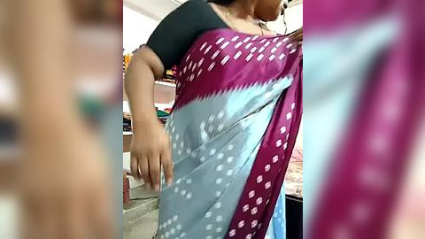 Media: Video of a woman in a maroon and light blue sari with white polka dots, wearing a black sleeveless top, standing in a cluttered room with shelves and clothes in the background.