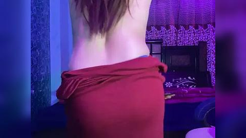 Media: Video of a woman with long brown hair, topless, wearing a red skirt, in a dimly lit room with purple and blue lighting, decorated with a patterned curtain and wooden furniture.