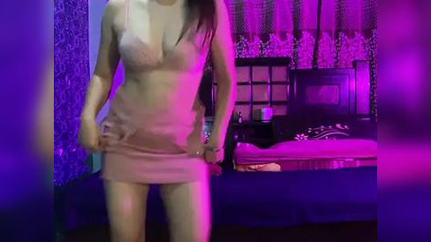 Media: Video of a woman in a sheer, nude-colored mini-dress, standing in a dimly-lit bedroom with purple lighting and a dark wooden bed.