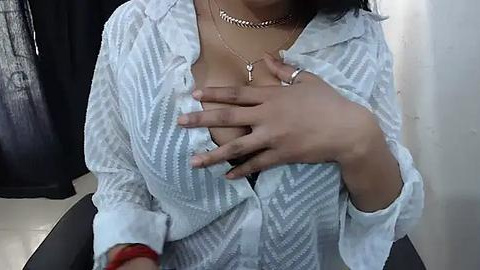 Media: A video of a woman with medium brown skin, wearing a sheer, light blue striped blouse, exposing cleavage. She is in a casual setting, with a black curtain in the background.