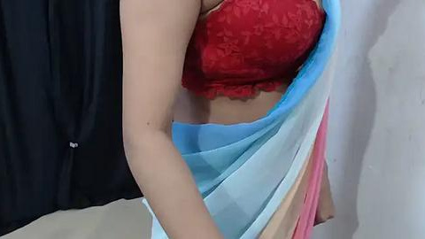 Media: Video of a woman's midsection, showcasing a red lace bra and a sheer blue and pink sari partially covering her midriff, against a plain white wall and black curtains in the background.