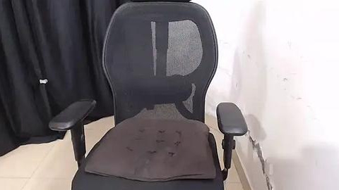 Media: Video of a black ergonomic office chair with mesh backrest and armrests, placed in a room with black curtains and white walls.