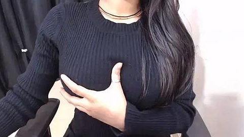 Media: Video of a woman with long, straight black hair, wearing a black ribbed sweater and a black choker necklace, cupping her left breast. Background features a white wall and a black garment on a hanger.