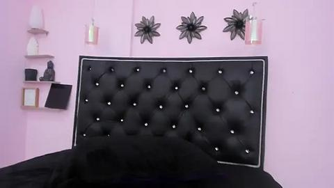 Media: A video of a minimalist, pink-walled bedroom with a black, tufted headboard adorned with small, silver buttons. Decorative black flower sculptures hang above the headboard, and a white lamp sits on a shelf to the left.