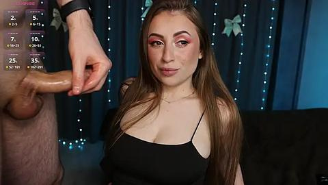 Media: Video of a fair-skinned woman with long brown hair, wearing a black top, smiling while holding a circumcised penis, in a dimly lit room with blue fairy lights.