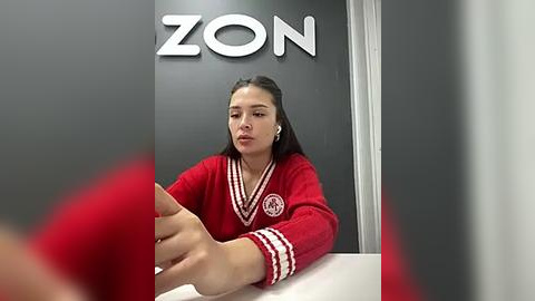 Media: Video of a woman with long black hair, wearing a red and white sweater, seated at a desk in a modern office with a black wall featuring the \"ZON\" logo.