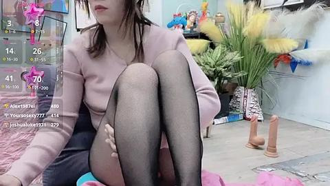 Media: Video of a woman in a light pink top, sitting cross-legged with black stockings, in a colorful, cluttered room with potted plants and toys.