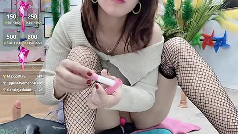 Media: Video of an Asian woman with fair skin, brown hair, and fishnet stockings, applying a pink vibrator to her vagina, wearing a cream sweater. Background includes a potted plant and colorful decorations.