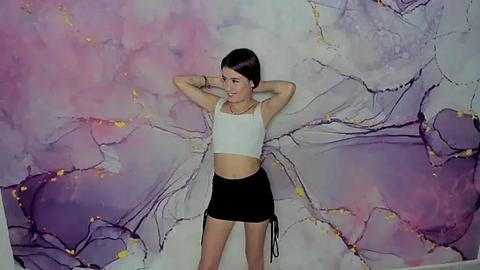 Media: Video of a young Asian woman with short black hair, wearing a white crop top and black shorts, posing confidently against a colorful, abstract marble-patterned wall.