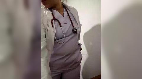 Media: Video of a woman in a white lab coat and light blue scrubs with a stethoscope, standing in a dimly lit, clinical setting.