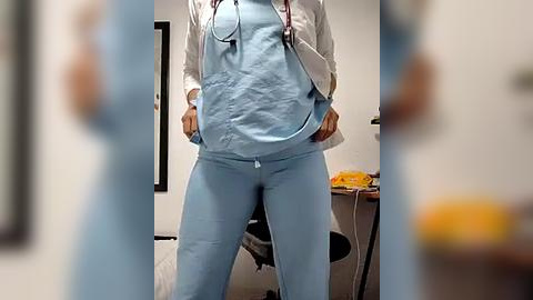 Media: A video of a woman in light blue scrubs and a white lab coat, hands in pockets, standing in a brightly lit room with a blurred background.