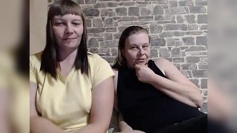 Media: Video of two women with light skin, one in a yellow top, the other in a black sleeveless top, seated against a rustic brick wall, appearing contemplative.