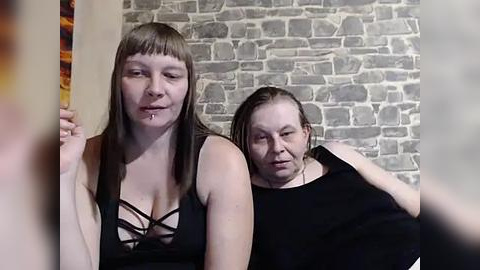 Media: Video of two women with pale skin and straight hair, one wearing a black bra, the other a black shirt, in a room with a stone wall.