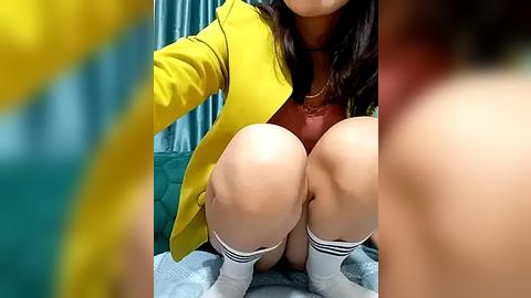 Media: A video of a woman with long dark hair, wearing a yellow blazer over a red top, sitting with knees up, legs in white socks, blurred background.