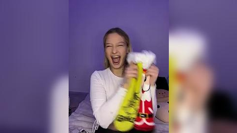 Media: Video of a joyful, fair-skinned woman with blonde hair, wearing a white shirt, holding a yellow-and-red toy gun, laughing in a dimly-lit room.