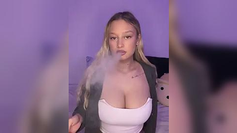 Media: Video of a young, light-skinned woman with long blonde hair, wearing a white tank top and grey blazer, smoking a cigarette. She has a tattoo on her right shoulder. The background is a dimly lit bedroom with a plush teddy bear.