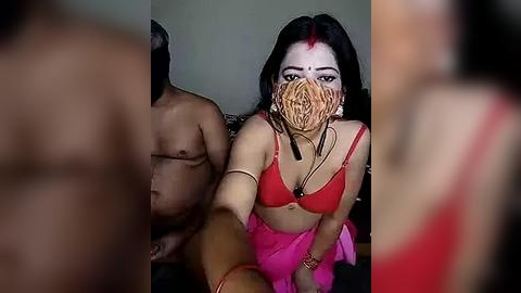 Media: Video: A woman with black hair, red forehead mark, and red mask, wearing a red bra, pink skirt, and arm bracelet, sits with a naked man behind her.