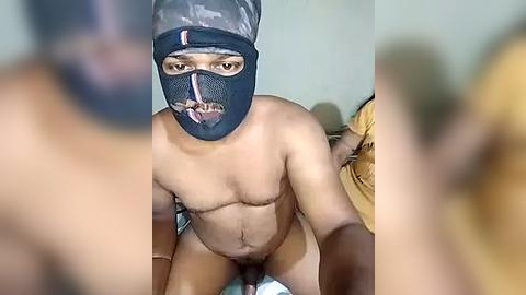 Media: A video shows a shirtless, muscular, light-skinned man with a black mask and a yellow and black camouflage hat, kneeling on a bed. The background is blurred, focusing on the man's torso and face.