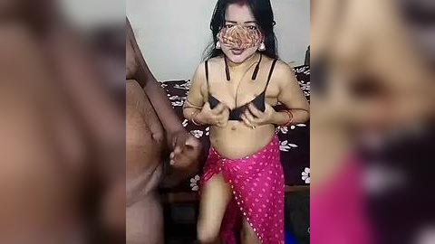 Media: A blurry video of an Asian woman with medium skin tone, long black hair, and medium-sized breasts, wearing a black bra and a pink sarong, holding a man's erect penis.