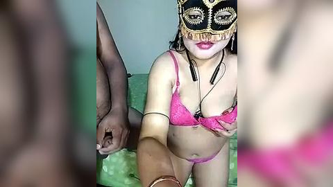 Media: A video shows a light-skinned woman with medium-length hair, wearing a black masquerade mask, pink lace lingerie, and black choker, holding a brown man's hand, on a green bed.