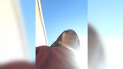 Media: Video of a young girl with medium brown skin, wearing a maroon sweater, standing against a bright blue sky, with a blurry foreground.