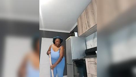 Media: Video of a Black woman with short hair, wearing a light blue tank top and pants, cleaning a modern kitchen with stainless steel appliances, wooden cabinets, and a white door.