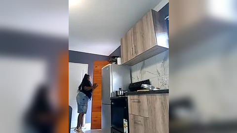 Media: A video shows a woman in a black top and denim shorts, standing in a modern kitchen with light wood cabinets, stainless steel appliances, and a gray marble backsplash.