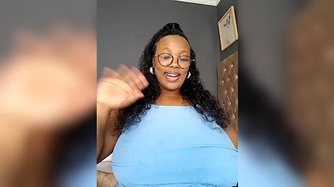 Media: Video of a smiling, light-skinned Black woman with curly hair and glasses, wearing a light blue top, sitting on a brown tufted leather sofa against a gray wall.