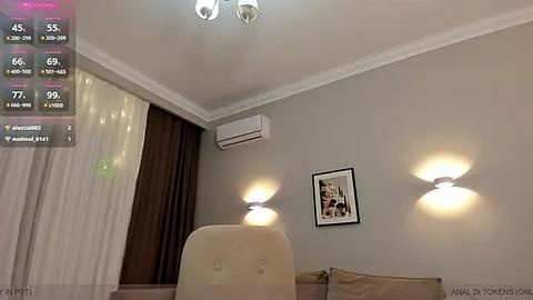 Media: Video of a modern, minimalist living room with beige walls, a beige sofa, white curtains, wall-mounted air conditioner, two wall-mounted sconces, and framed artwork.