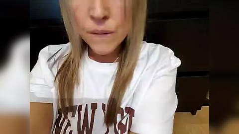 Media: Video of a young woman with long, straight blonde hair, wearing a white \"Wolverines\" T-shirt, sitting indoors. Her face is partially obscured by shadows.