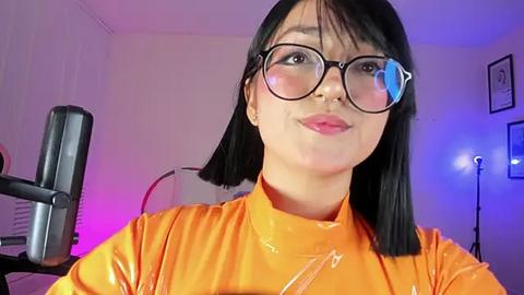 Media: Video of a young East Asian woman with straight black hair, wearing large round glasses and an orange latex dress, in a dimly lit room with a black gaming chair and blue and purple lighting.