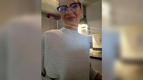 Media: Video of a smiling person with glasses, wearing a ribbed light gray sweater, standing in a kitchen with white cabinets and a microwave.
