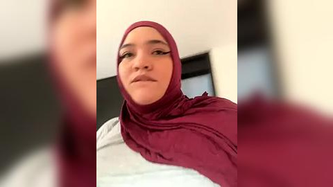 Media: Video of a young woman with medium skin tone, wearing a red hijab and white top, indoors, with a blurred background.